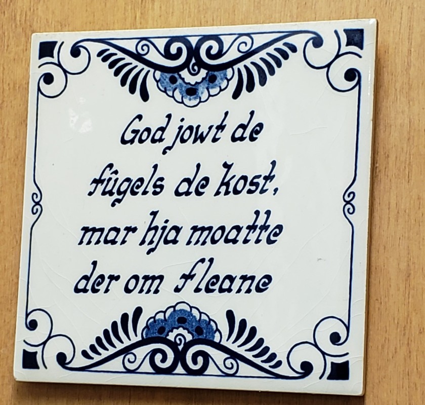 White tile with blue embellishments, and text in blue reading "God jowt de fugels de kost, mar hja moatte der om fleane"
